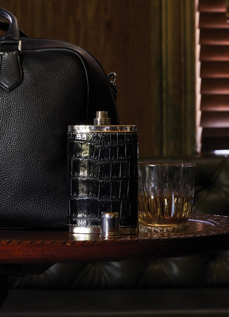 Black crocodile covered hip flask on table next to a glass - Darby Scott